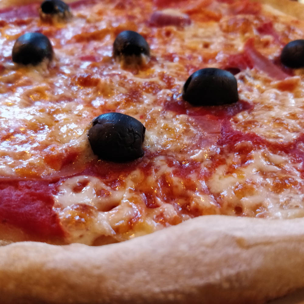 Nahaufnahme einer gebackenen Pizza. Im Vordergrund ein Stück Kruste, dahinter Tomatensauce, mit viel Käse, Schinken und einigen schwarzen Oliven.